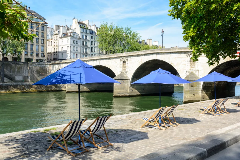 Three blue parasols with blue and white striped deck chairs in the sun on the bank of the river Seine with typical parisian bridge and buildings in the background.